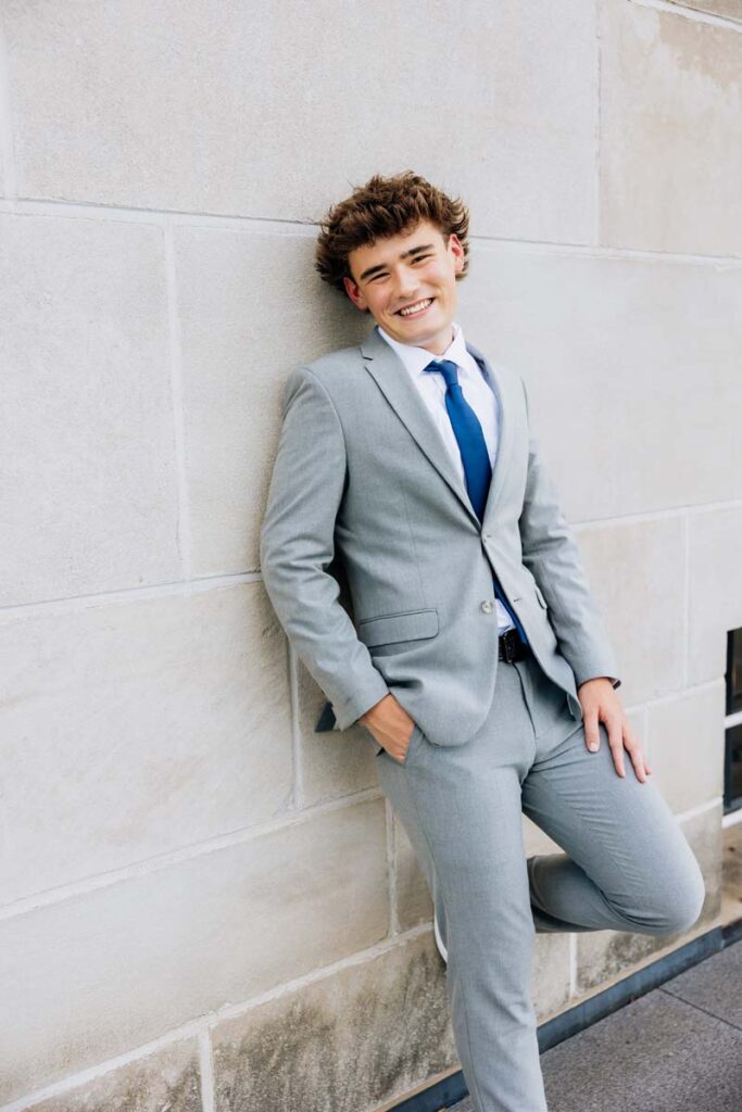 Senior boy in gray suit and blue tie leaning against modern wall Lincoln Nebraska state capital 